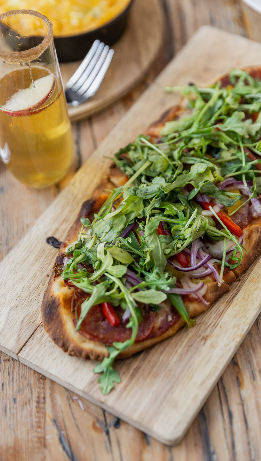 Wood-fired flatbread topped with arugula, salami, red onion, and peppers on a wooden board